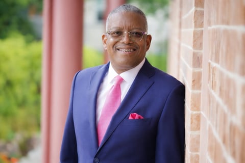 Professional headshot of a man in a navy blue suit, pink tie, and glasses, smiling in front of a brick wall with red columns