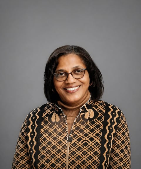 Professional headshot of a smiling woman with black shoulder-length hair and glasses, wearing a patterned black and gold jacket against a gray background