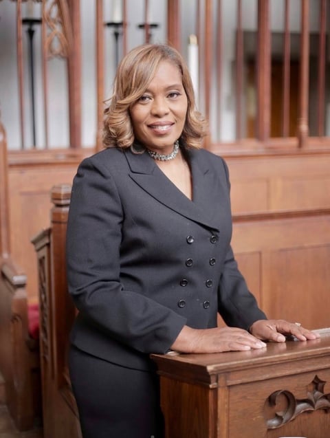 Professional woman in dark blazer standing at wooden desk in courtroom setting, wearing pearl necklace and smiling at camera